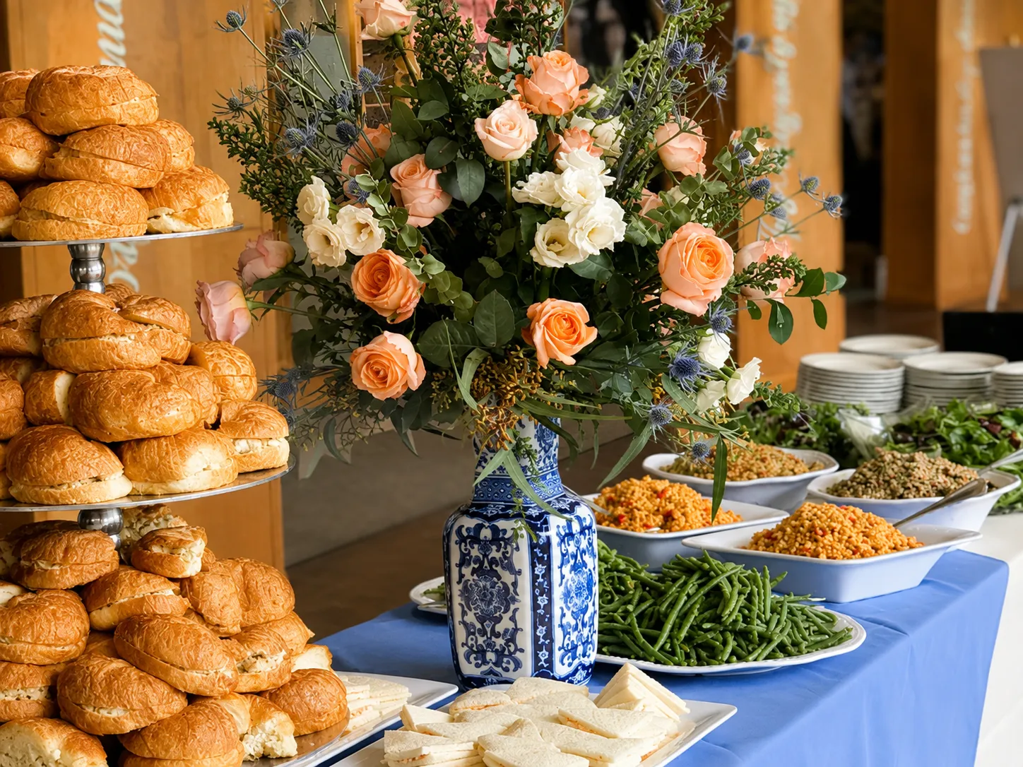 A buffet table with a blue cloth holding a tiered silver stand stacked with croissant sandwiches, a tall blue and white porcelain vase of peach roses, cream lisianthus, blue thistle and eucalyptus, platters of green beans, tomato rice, lentil salad and greens, and a white tray of finger sandwiches.