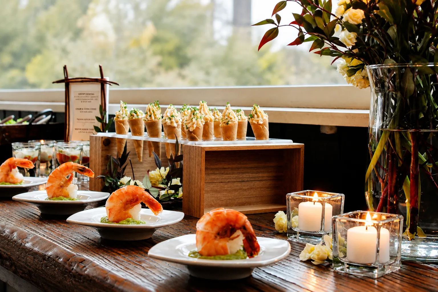 A wooden display table by a window with a row of savory cones piped with crab and corn filling on a riser, small white plates of jumbo shrimp cocktail over avocado, layered shrimp verrines in glasses, votive candles in square holders, and a tall glass vase of cream roses and red-tipped foliage.