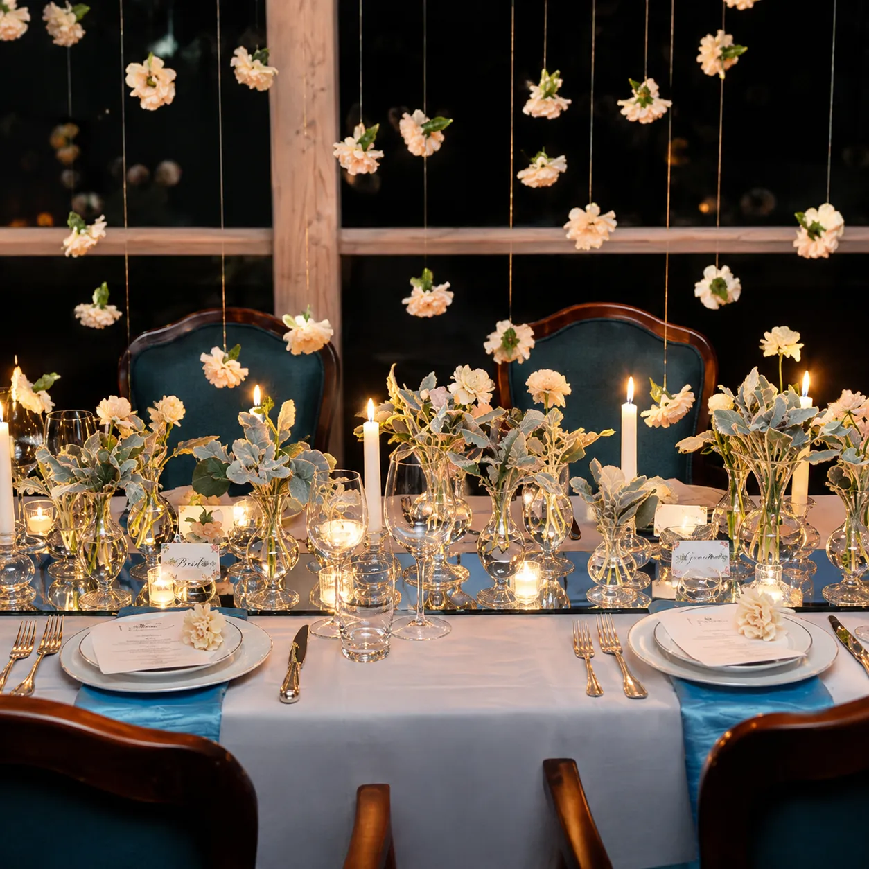 A candlelit wedding head table with a white cloth and blue runner, gold flatware, menu cards on gilded plates, clear vases of peach zinnias and dusty green foliage, tapered candles, and peach blooms hanging on gold threads above teal upholstered chairs.