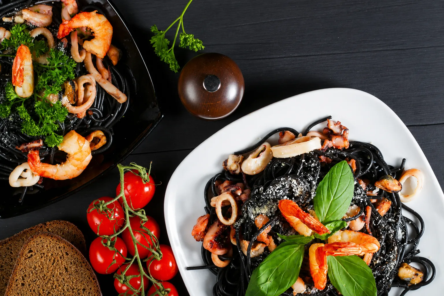 Overhead view of two servings of squid ink spaghetti with shrimp, calamari rings, octopus, mussels and grated cheese, one on a black dish with parsley and one on a white plate with basil, beside vine cherry tomatoes, slices of dark rye bread and a wooden pepper mill on a black wood table.