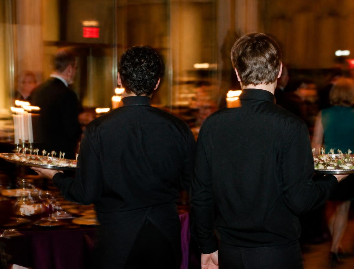 Service staff in black uniforms carrying trays of canapes through a candlelit reception.
